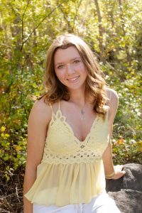 Young woman in a yellow lace dress smiling outdoors in a sunlit forest setting.