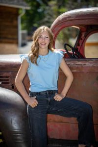 Young woman in a light blue top posing with a rusty vintage truck outdoors, hands on hips and smiling